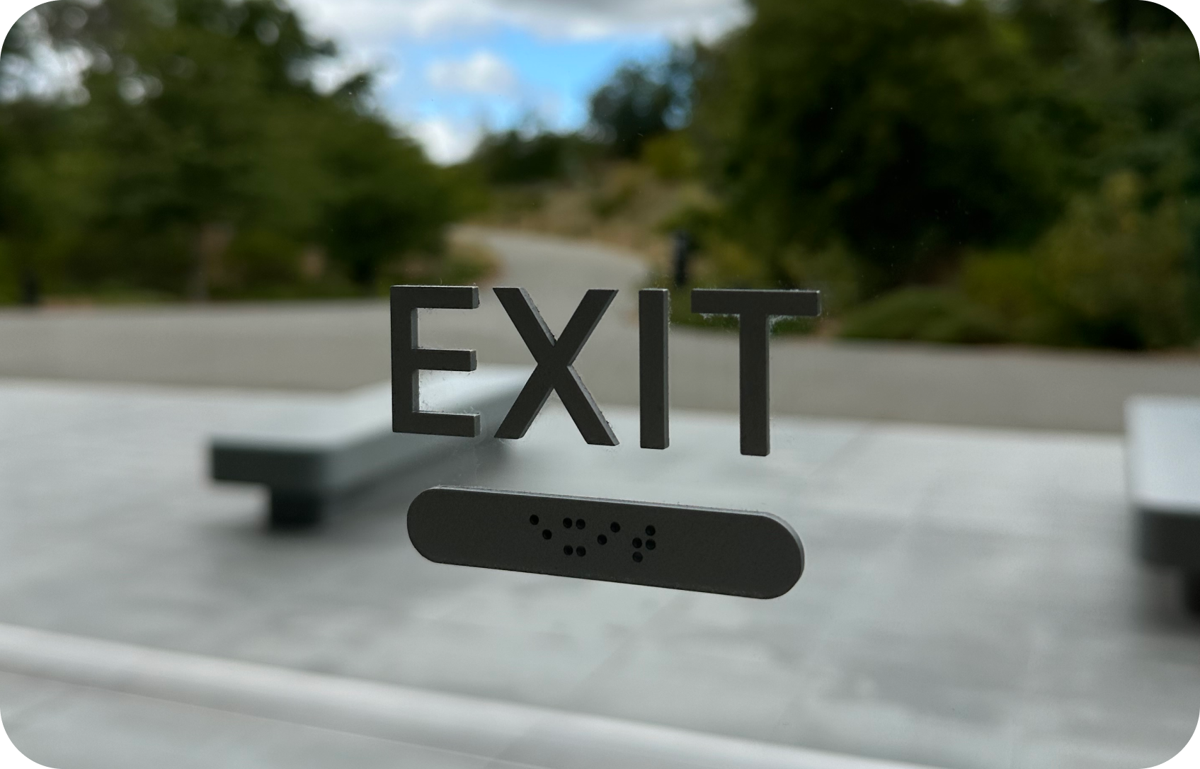The word EXIT with the corresponding braille text under it glued to a glass wall. A lush park with a path leading outward, as-if to signify the forward-only path that awaits one’s exit
